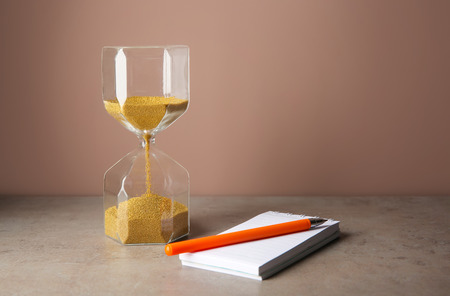 Hourglass with notebook and pen on table against color background. Time management conceptの写真素材