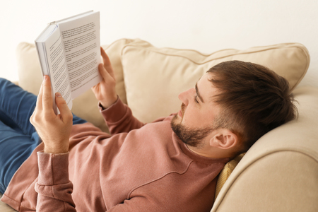 Young man reading book while lying on sofa at homeの写真素材