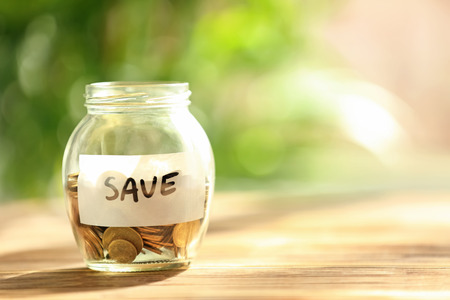 Coins in glass jar with text SAVE on table against blurred background. Concept of savings