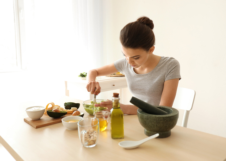 Young woman making nourishing mask with avocado in kitchenの写真素材