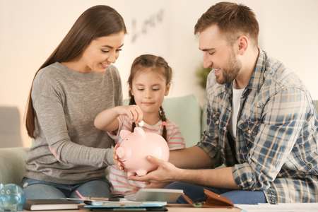 Little girl with her parents sitting on sofa and putting coin into piggy bank indoors. Money savings conceptの写真素材