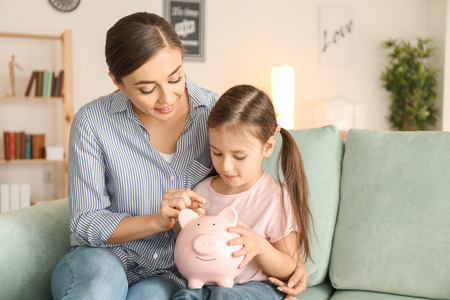 Little girl with her mother sitting on sofa and putting coin into piggy bank. Money savings conceptの写真素材
