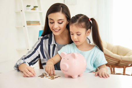 Little girl with her mother sitting at table and putting coins into piggy bank indoors. Money savings conceptの写真素材