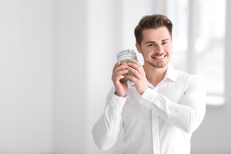 Young businessman holding glass jar with coins indoors. Money savings conceptの写真素材