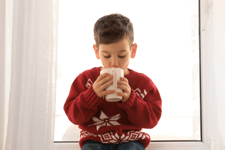 Cute little boy with cup of hot cocoa drink near windowの写真素材