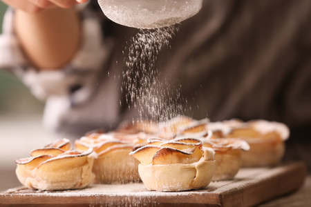Woman sprinkling apple roses from puff pastry with sugar powder in kitchen, closeupの写真素材