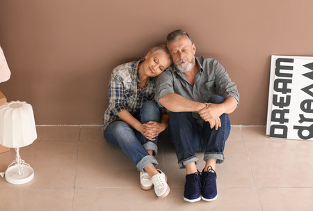 Happy mature couple with belongings sitting on floor indoors. Moving into new houseの写真素材