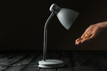 Woman holding coins near lamp on wooden table. Electricity saving conceptの写真素材