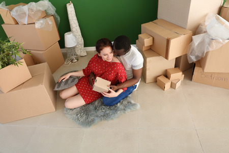 Interracial couple sitting on floor with boxes indoors. Moving into new house