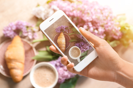 Woman taking photo of croissant with coffee and blossoming lilacの写真素材