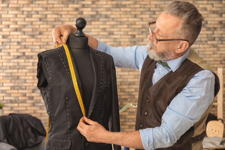 Mature tailor taking measurements of jacket on mannequin in atelier