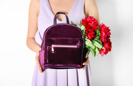 Young woman holding backpack with beautiful peonies on light background