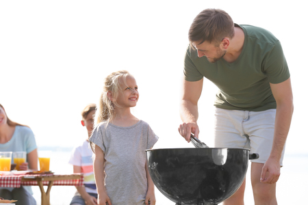 Young man with daughter cooking tasty food on barbecue grill outdoors. Family picnicの写真素材