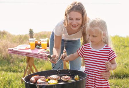 Young woman with daughter cooking meat, sausages and vegetables on barbecue grill outdoorsの写真素材