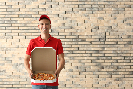 Young man holding box with tasty pizza against brick wall. Food delivery serviceの写真素材