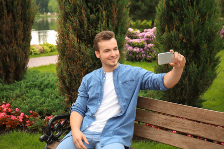 Young man taking selfie while sitting on wooden bench in parkの写真素材
