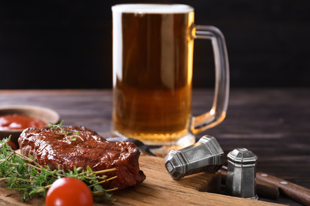 Wooden board with tasty grilled steak and mug of beer on tableの写真素材