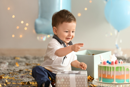 Cute little boy with gift box and birthday cake sitting on carpet in roomの写真素材