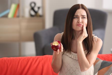 Young woman with sensitive teeth and apple on sofa at homeの写真素材
