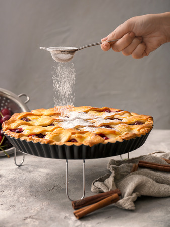 Woman sprinkling sugar powder over sweet homemade cherry pie in kitchenの写真素材
