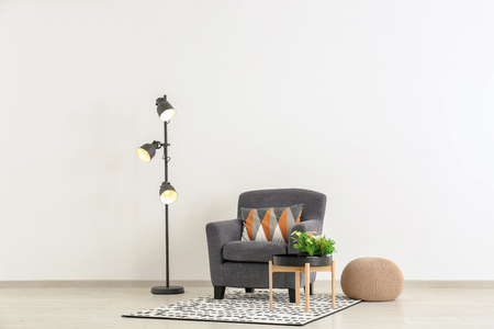 Comfortable armchair with table, pouf and floor lamp near light wallの写真素材