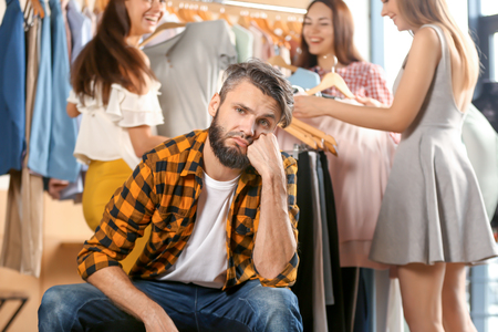 Annoyed young man waiting for his girlfriend and her friends while they choosing clothes in shopの写真素材