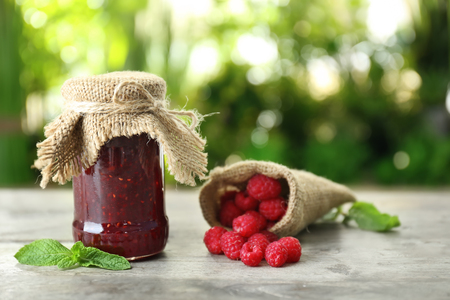 Glass jar with tasty raspberry jam on table against blurred background