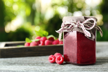 Glass jar with tasty raspberry jam on table against blurred backgroundの写真素材
