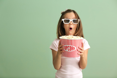Emotional little girl with cup of popcorn watching movie on color backgroundの写真素材