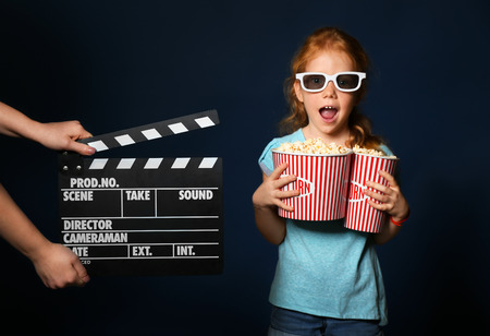 Female hands with clapper board and cute girl holding cups of popcorn on dark backgroundの写真素材