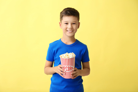 Cute little boy with cup of popcorn on color backgroundの写真素材