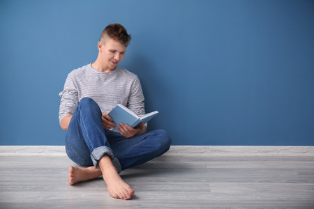Portrait of handsome young man reading book while sitting on floor near color wallの写真素材