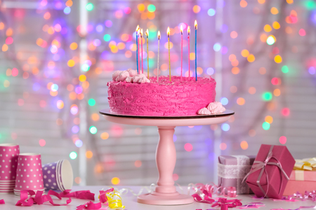 Stand with delicious birthday cake and burning candles on table against blurred lightsの写真素材