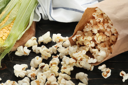 Paper bag with scattered delicious popcorn and cob on wooden background, closeup