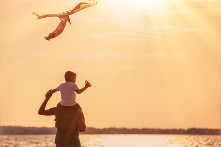 Happy father and son flying kite near river at sunsetの写真素材