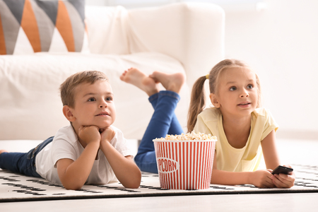 Cute children eating popcorn while watching TV at homeの写真素材