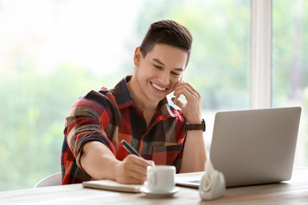 Young freelancer talking by mobile phone while working in cafeの写真素材