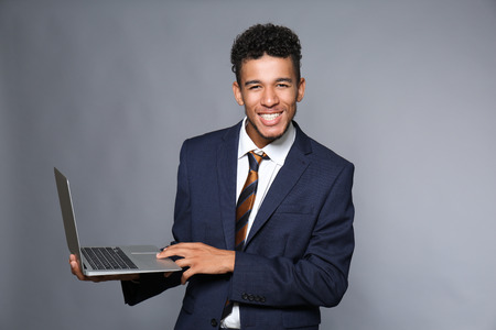 Handsome African-American businessman with laptop on grey background