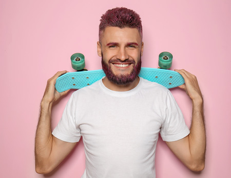 Portrait of handsome man with dyed hair and beard holding skateboard on color backgroundの写真素材