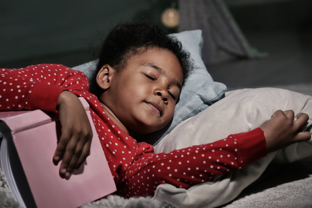 Little African-American girl with book sleeping in bedの写真素材