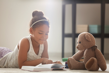 Little African-American girl reading book while lying on floor at homeの写真素材