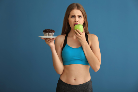 Young woman in sportswear choosing between cake and apple on color backgroundの写真素材