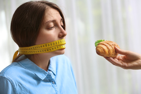 Offering of tasty croissant sandwich to young woman with measuring tape around her mouth on light background. Diet conceptの写真素材
