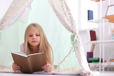 Cute little girl reading book in hovel at homeの写真素材