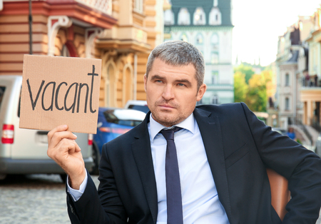 Businessman holding piece of cardboard with word VACANT while sitting in office armchair outdoorsの写真素材
