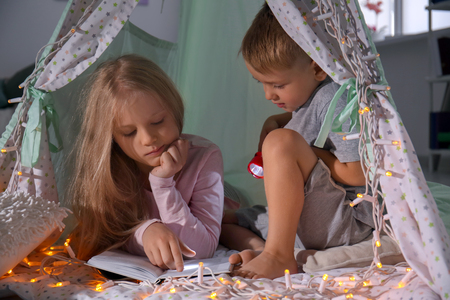 Cute little children reading book in hovel at homeの写真素材