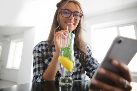 Young woman with mobile phone drinking fresh lemonade at homeの写真素材