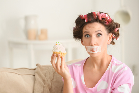 Emotional woman with taped mouth holding tasty tartlet at home. Diet conceptの写真素材