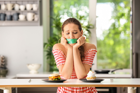 Emotional woman with measuring tape around her mouth and unhealthy food in kitchen. Diet conceptの写真素材
