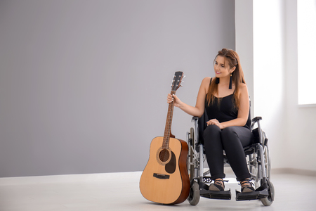 Beautiful young musician with guitar sitting in wheelchair indoorsの写真素材